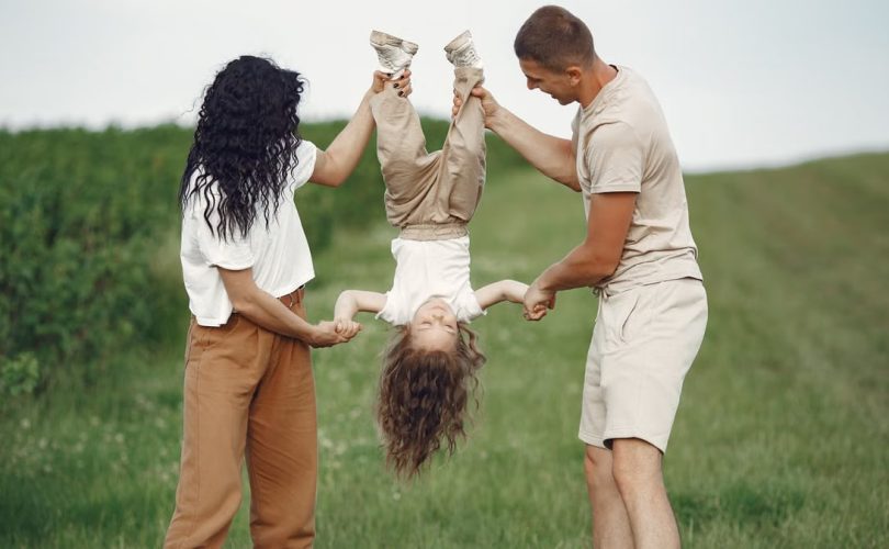 A joyful family enjoys outdoor play, lifting their child upside down in a green field.